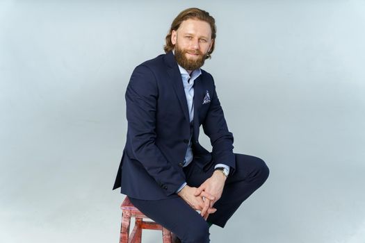 a handsome brutal bearded and long-haired man, a stylish businessman sitting on a chair with a white background. High quality photo