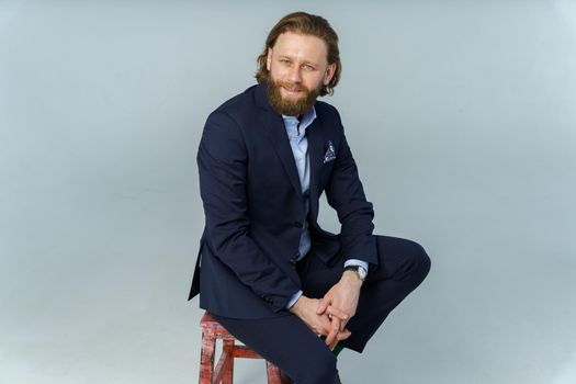 a handsome brutal bearded and long-haired man, a stylish businessman sitting on a chair with a white background. High quality photo