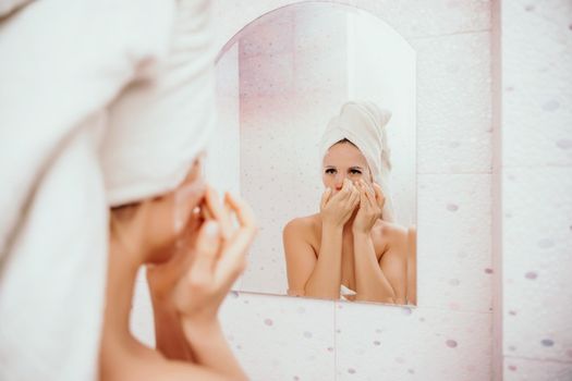 Young beautiful woman using a moisturizing facial mask after taking a bath. Pretty attractive girl in a towel on her head stands in front of a mirror in a home bathroom. Daily hygiene and skin care.