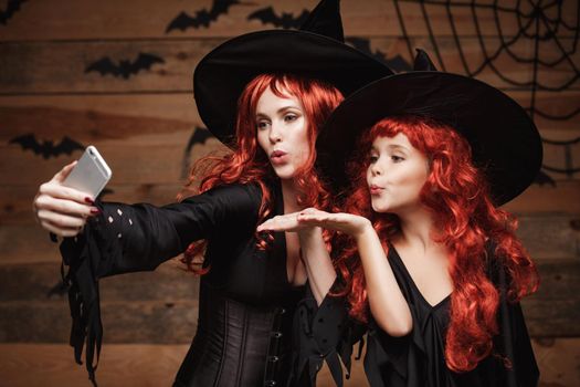 Halloween Concept - Beautiful caucasian mother and her daughter with long red hair in witch costumes taking a selfie with smartphone ready for celebrating Halloween