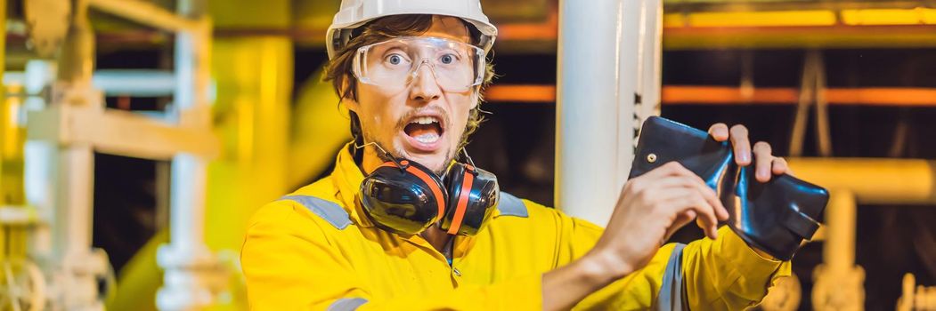 BANNER, LONG FORMAT Young man in a yellow work uniform, glasses and helmet in industrial environment,oil Platform or liquefied gas plant. looking into his empty wallet. Do not pay salary.