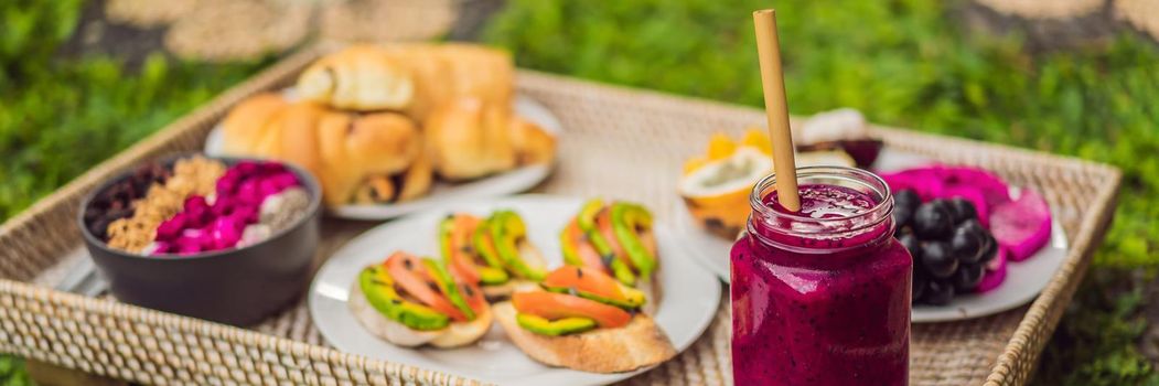 BANNER, LONG FORMAT Breakfast on a tray with fruit, buns, avocado sandwiches, smoothie bowl standing on the grass.