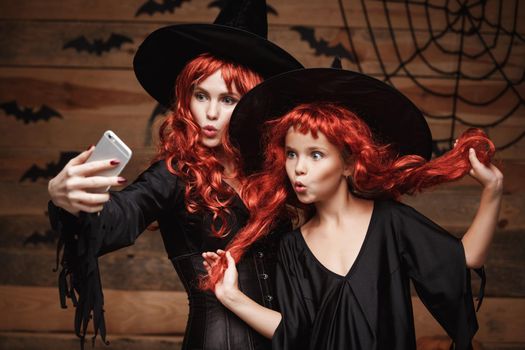 Halloween Concept - Beautiful caucasian mother and her daughter with long red hair in witch costumes taking a selfie with smartphone ready for celebrating Halloween