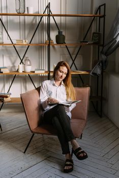 Beautiful lady sitting in a armchair and reading a book. Young serene woman relaxing at home and reading a book in the sun light.