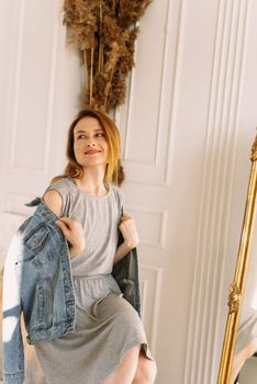 Cozy home atmosphere. Stylish redhead woman in gray dress and jeans sits on a chair at home next to a mirror and wheat. Portrait beautiful young woman happy smile relax in living room interior.
