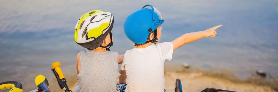 Two boys sit on the shore of the lake after riding a bike and scooter. BANNER, LONG FORMAT