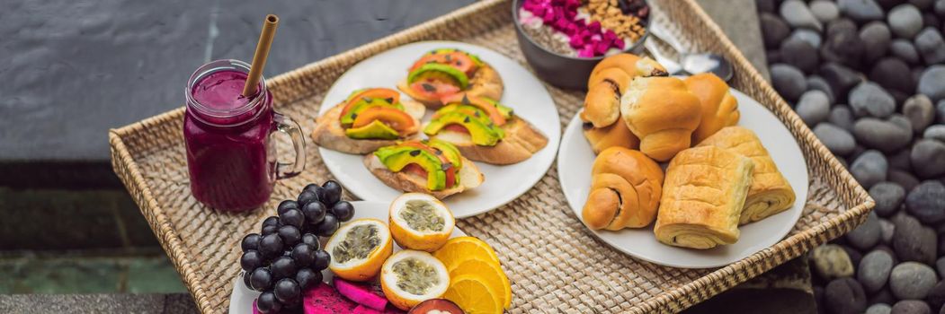 Breakfast on a tray with fruit, buns, avocado sandwiches, smoothie bowl by the pool. Summer healthy diet, vegan breakfast. Tasty vacation concept. BANNER, LONG FORMAT