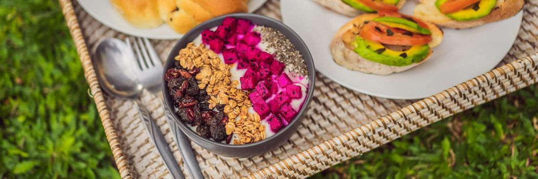 BANNER, LONG FORMAT Breakfast on a tray with fruit, buns, avocado sandwiches, smoothie bowl standing on the grass.