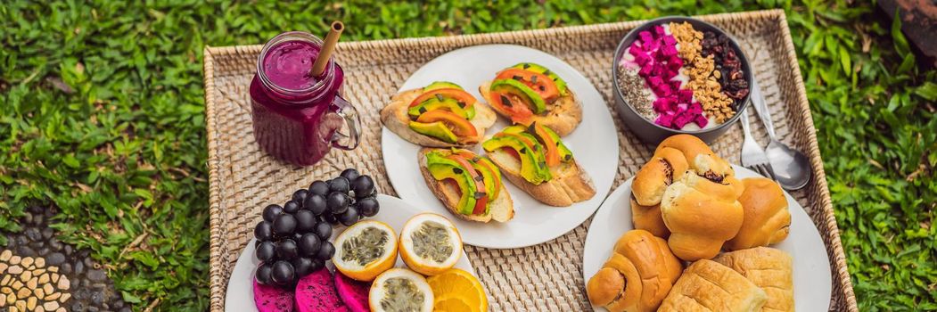 BANNER, LONG FORMAT Breakfast on a tray with fruit, buns, avocado sandwiches, smoothie bowl standing on the grass.