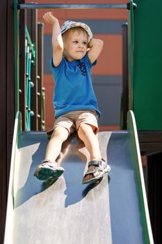 Cheerful little boy in a blue T-shirt and beige shorts rolls down a metal slide in city park. Summer active sport leisure for kids. Daycare for young kid. Baby on play ground. Outdoor fun in summer.