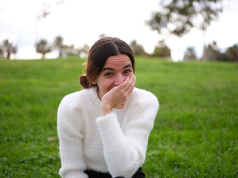 Front view of a happy caucasian young woman in the park looking at the camera covering her mouth with her hand while laughing. Concept of emotions.