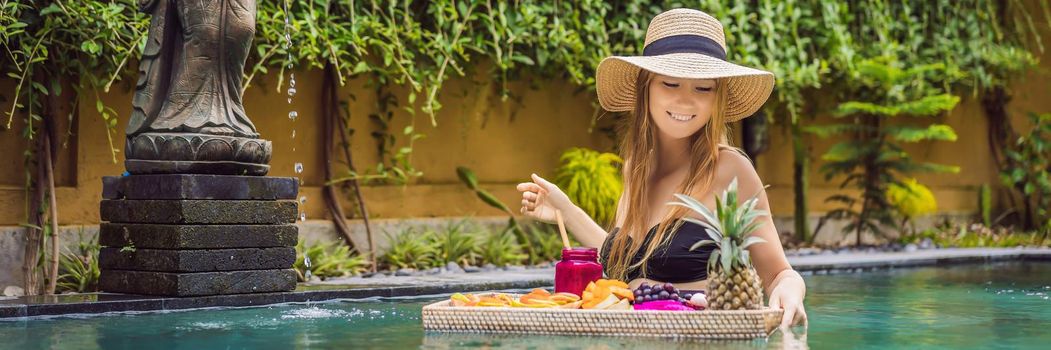 BANNER, LONG FORMAT Breakfast tray in swimming pool, floating breakfast in luxury hotel. Girl relaxing in the pool drinking smoothies and eating fruit plate, smoothie bowl by the hotel pool. Exotic summer diet. Tropical beach lifestyle. Bali Trend.