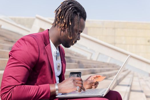 elegant african man sitting outdoors paying with his credit card for the online purchase done on his laptop computer, technology and online shopping concept