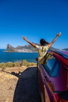 woman outside car window road trip with hands up, car at Chapmans Peak Drive in Cape Town South Africa looking out over the ocean.