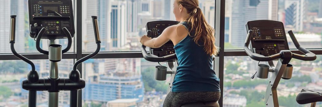 Young woman on a stationary bike in a gym on a big city background. BANNER, LONG FORMAT