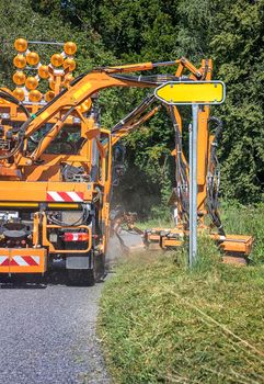 Tractor with a mechanical mower mowing grass on the side of the asphalt road. Road services are engaged in landscaping around roads. Vertical view