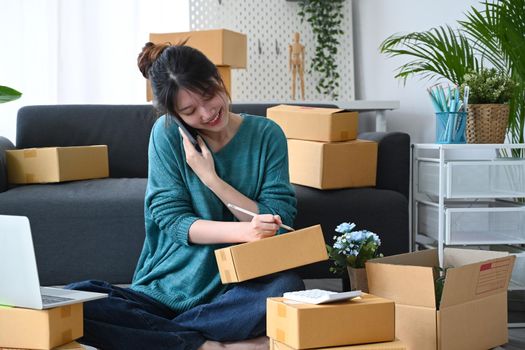 Smiling asian woman online seller receive order on mobile phone and preparing parcel boxes of product for delivery.