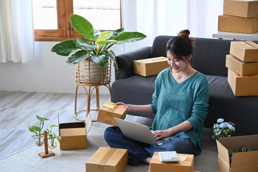 Smiling young woman using laptop and preparing parcel boxes of product for delivery to customers. E-commerce, Online selling concept.