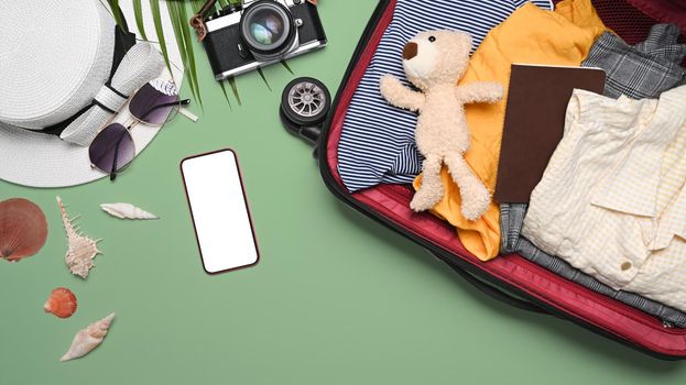 Open suitcase with clothes, smart phone, camera, hat and sunglasses on green background. Packed for journey.