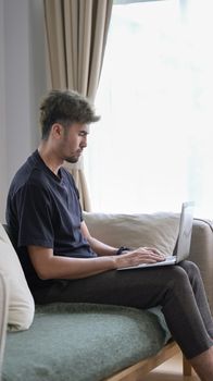 Man in casual clothes using laptop computer, surfing internet while sitting on couch at home.