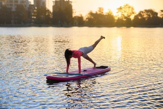 Healthy and fit woman balancing on sup board while doing yoga exercises. Caucasian brunette in sportswear having active workout on summer lake.
