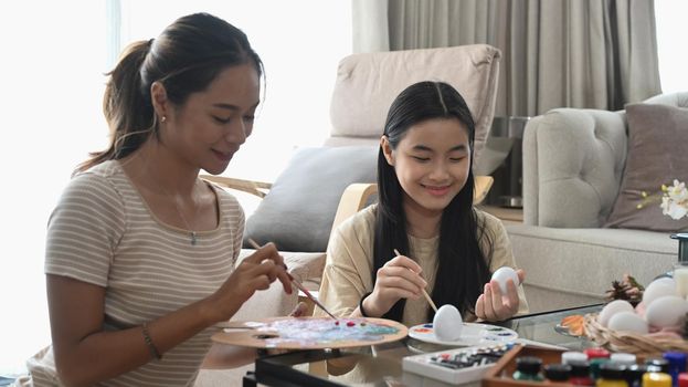 Mom and daughter are sitting in bright living room and coloring Easter eggs.