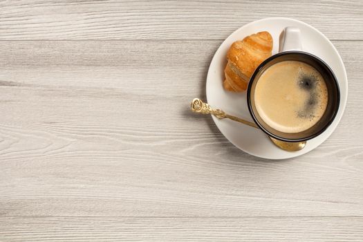 Top view of cup of black coffee with fresh croissant and spoon on saucer with copy space on grey wooden background