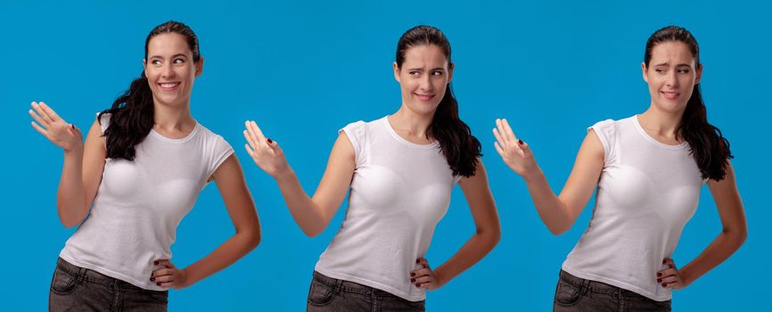 Close-up shot of a beautiful brunette girl in a white casual t-shirt smiling and posing against a blue studio background. She act like refusing someone. Set of a people sincere emotions. Copy space.