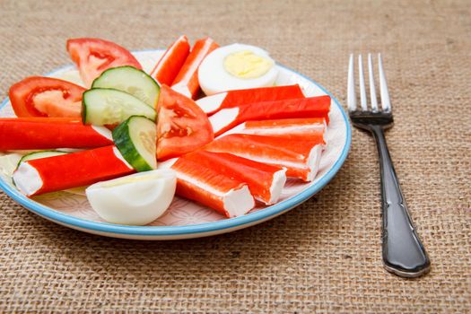 Plate with crab sticks, boiled egg and freshly sliced tomato and cucumber with fork on sackcloth
