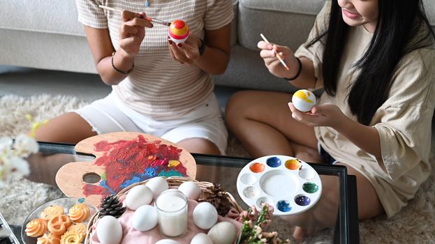 Smiling little girl and mother sitting floor in living room and painting Easter eggs together. Easter, holidays and people concept.