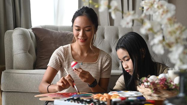 Happy mother and daughter enjoy painting eggs while preparing for Easter egg festival at home.