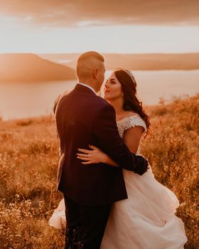 A loving couple wedding newlyweds in white dress and suit walk hug kissing whirl on tall grass in summer field on mountain above the river.style bride accessories on head crown makeup hairstyle