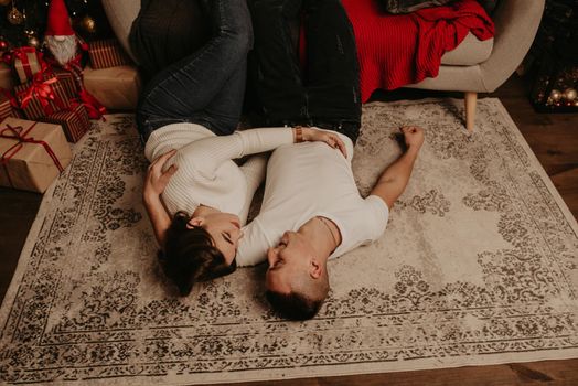 young couple man and woman lie on floor near couch with legs feet up hugging kissing in the bedroom near christmas tree. decorated house for New Year. Valentine's Day celebration
