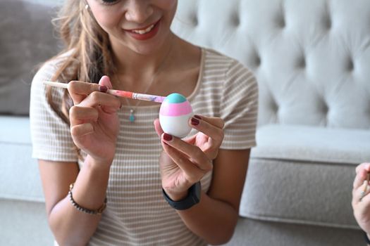 Smiling young woman sitting in living room and enjoy painting water colors on eggs for Easter festival.