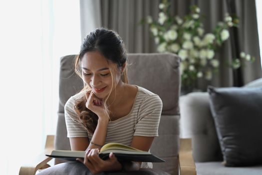 Happy millennial woman reading book while sitting on beige armchair in apartment.