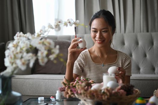 Beautiful young woman sitting in living room and painting egg for Easter festivity. Easter holidays concept.