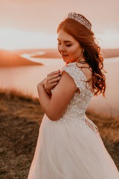 young brunette bride in white wedding dress with a crown on her head stands on cliff against the background of the river and islands After sunset.style fashionable women hairstyle makeup