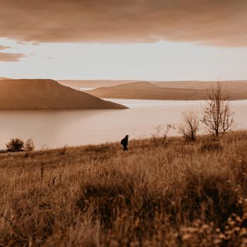 man traveler backpacker in casual clothes stands on mountain. sunset rays through clouds. panoramic view from cliff to river lake sea in distance silhouettes islands