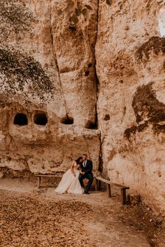 wedding couple in love man and woman sitting on a bench under rock monastery bakota in autumn forest against background of stone rocks. groom suit and bride dress with crown on head