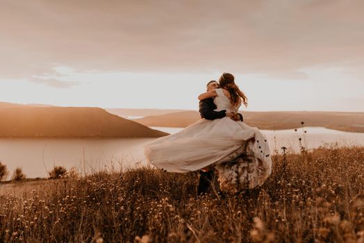 loving couple wedding newlyweds in white dress and suit hug kissing whirl on tall grass in summer field on mountain above the river. sunset.