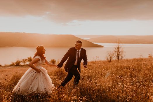 A loving couple wedding newlyweds in a white dress and a suit walk run smile happy on tall grass in the summer field on the mountain above the river. sunset.
