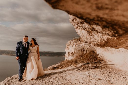 loving couple wedding newlyweds in white dress and suit walk in summer on mountain above river. sunset and sunrise. man and woman on rocks above cliff