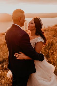 A loving couple wedding newlyweds in white dress and suit walk hug kissing whirl on tall grass in summer field on mountain above the river.style bride accessories on head crown makeup hairstyle