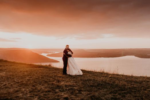 A loving couple wedding newlyweds walk in tsummer in the fall on mountain on cliff above the river. sunset. bride in a white dress with crown on her head. style fashionable women hairstyle makeup