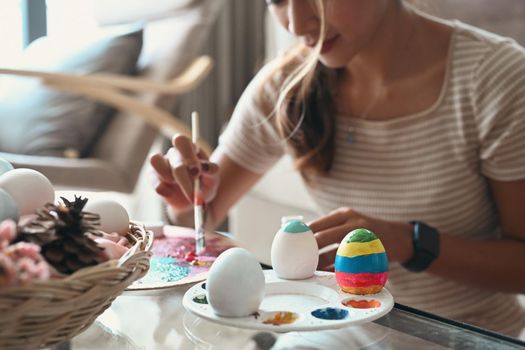 Pretty young woman holding paintbrush and painting egg, preparing for Easter.