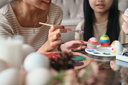 Happy young mother hands holding paintbrush helping her daughter painting Easter eggs. Easter, holidays and people concept.