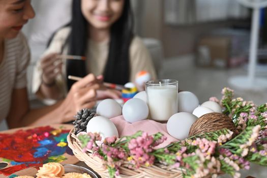 Wicker basket decorating with candle, eggs and flowers in living room with mother and daughter painting Easter egg in background.