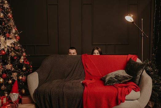 man and a woman hid behind couch. heads of a man and a woman stick out from behind sofa. decorated house for New Year. Christmas morning. apartment interior. Valentine's Day celebration