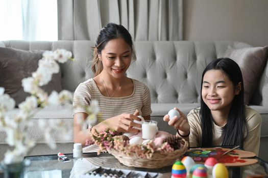 Cute asian girl and mother are arranging Easter eggs into wicker basket, preparing for Easter festival.