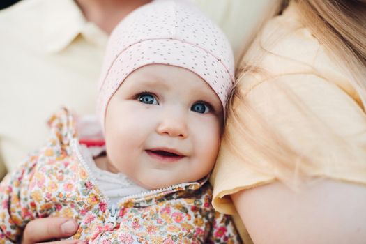 Close up of surprised baby's face with lightly opened mouth. Sweet lovely child in colorful clothes and hat leaning on mother's back. Father's hand holding infant while it looking at camera.